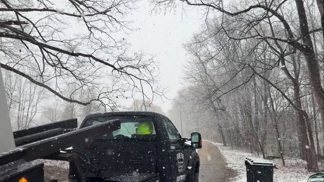 Pickup truck on a snowy, tree-lined road with snow falling and another vehicle behind it