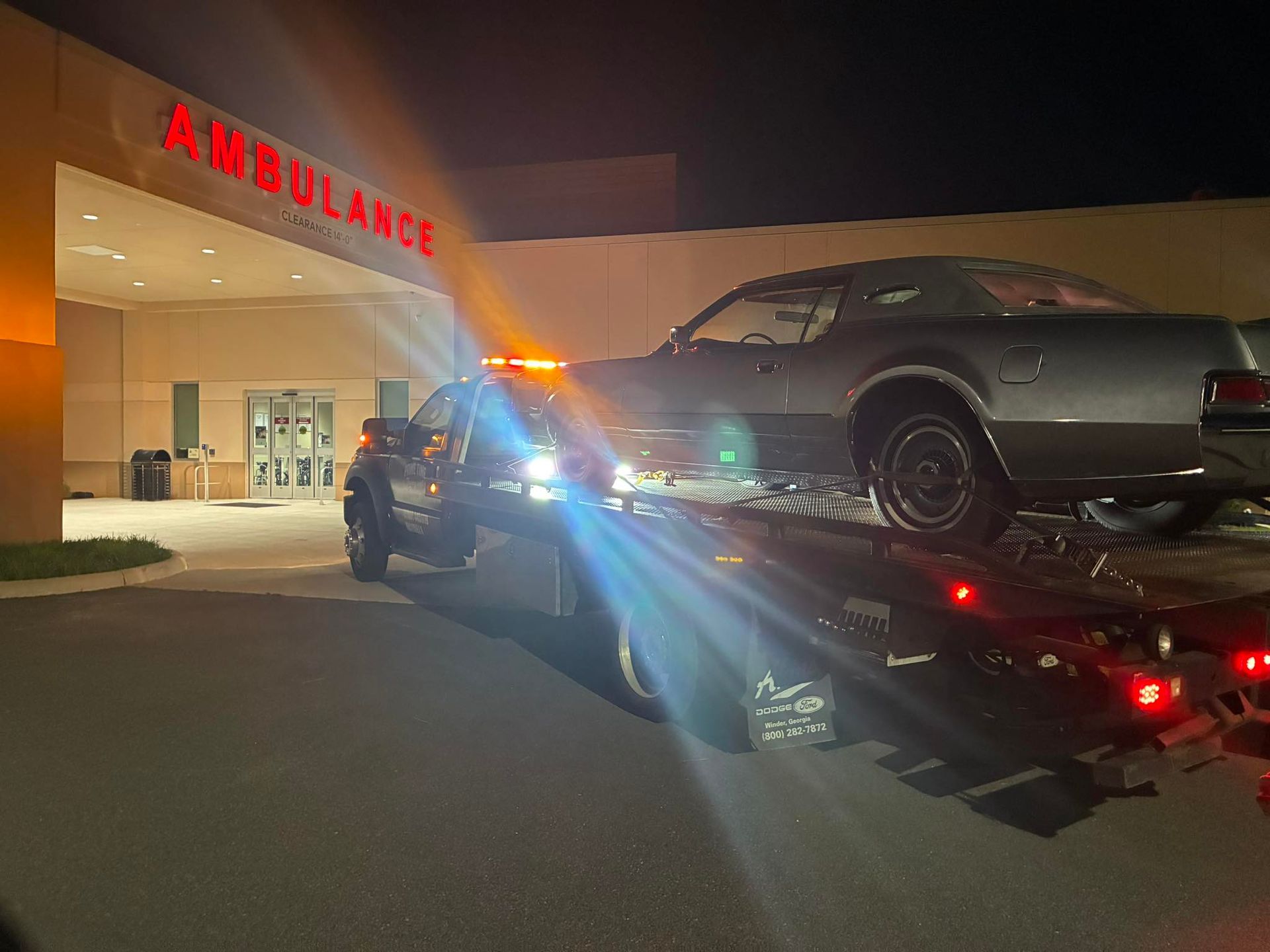 Ambulance at night with a car on a tow truck outside a lit building