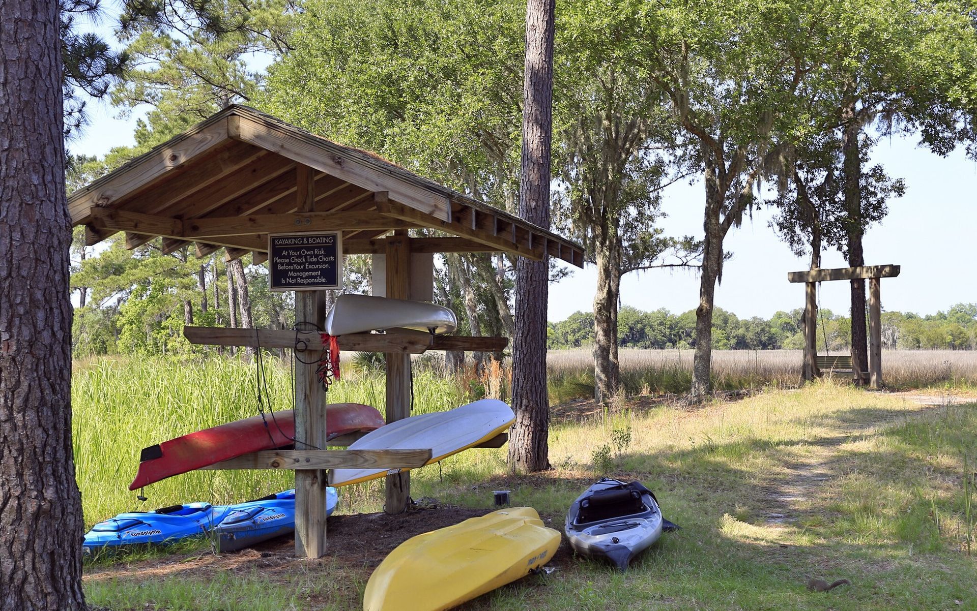 Kayak shelter with colorful kayaks on the ground under trees, beside a grassy field and riverbank at Grand Oaks at Ogeechee River in Savannah, GA.