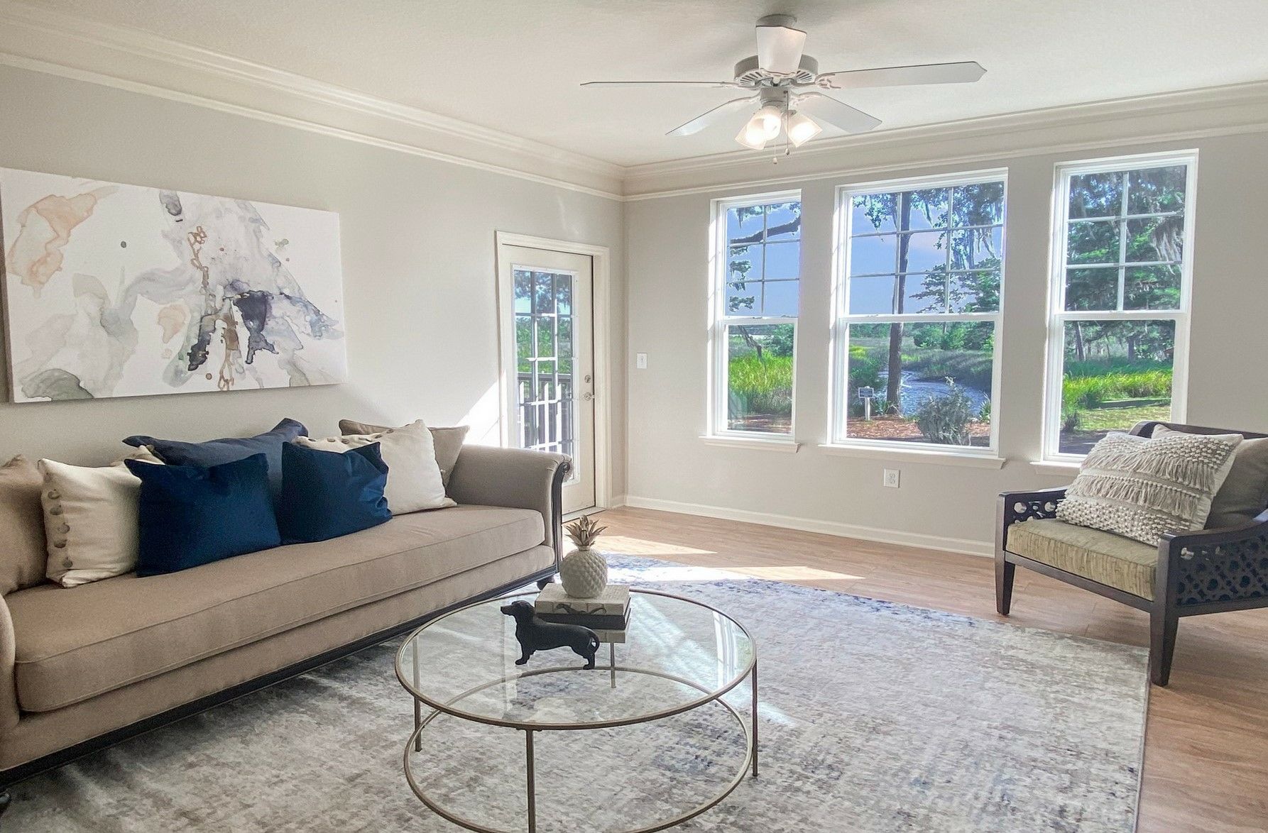 Bright living room with beige sofa, blue pillows, glass coffee table, and large windows letting in sunlight at Grand Oaks at Ogeechee River in Savannah, GA.