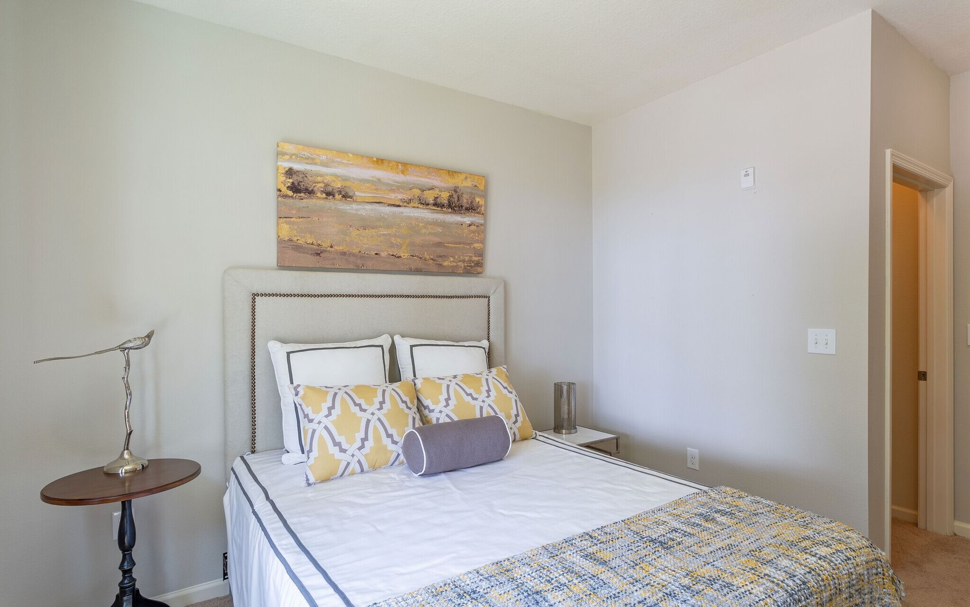 Small bedroom with a bed, patterned pillows, side table, and abstract wall art in neutral tones at Grand Oaks at Ogeechee River in Savannah, GA.