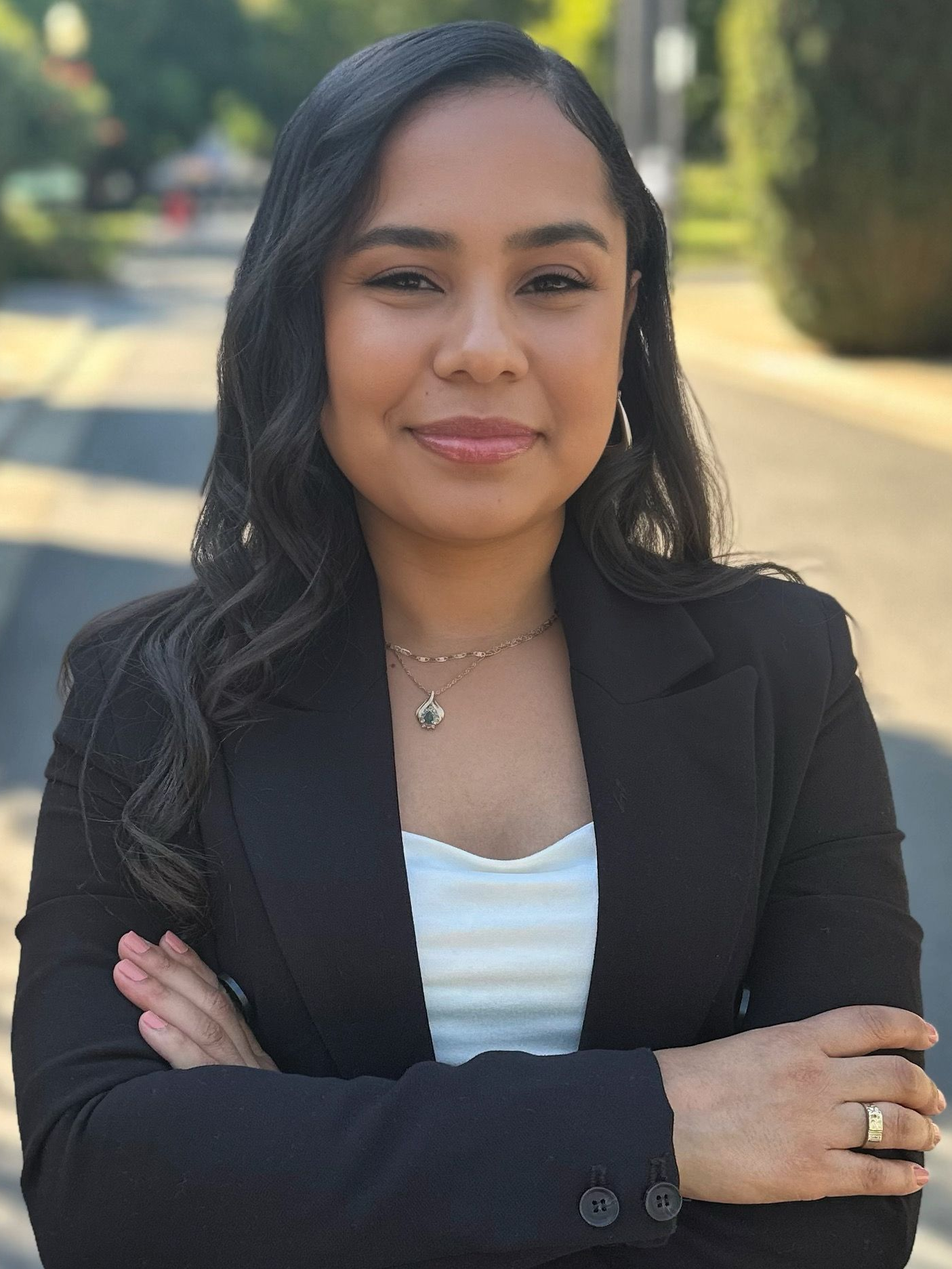 Woman in a black blazer standing outdoors with arms crossed, smiling in soft sunlight.