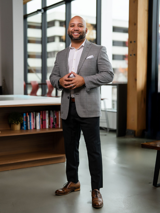 Man standing indoors in a gray blazer, smiling in a modern office lobby near bookshelves and windows
