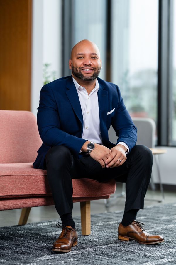 Man in navy blazer seated on a pink bench in a bright office lounge, smiling at the camera.