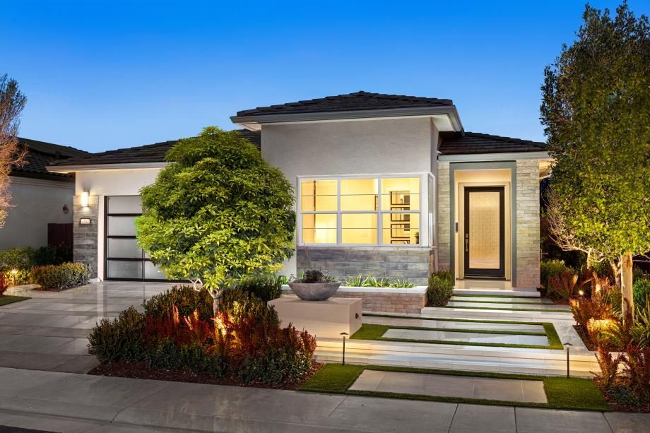 Modern single-story house with lit windows, front steps, and landscaped yard at dusk
