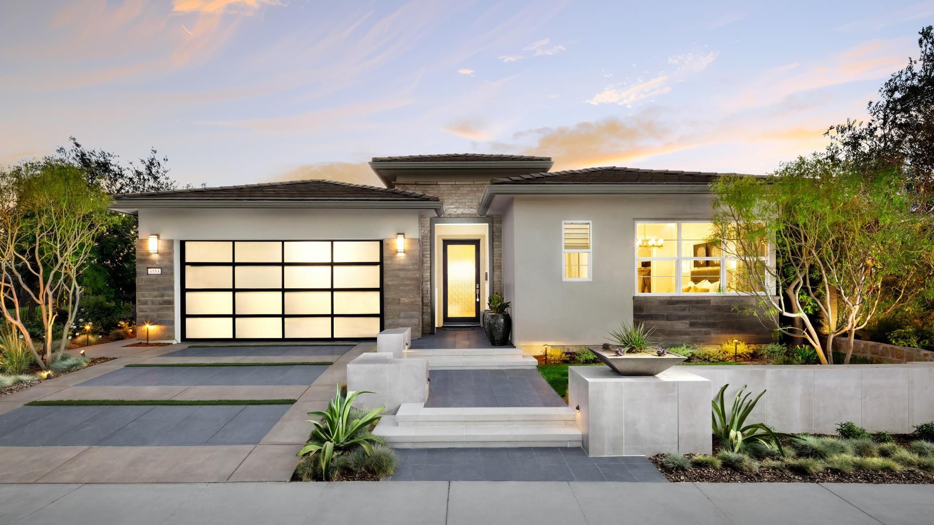 Modern single-story house at dusk with lit windows, driveway, and landscaped front yard