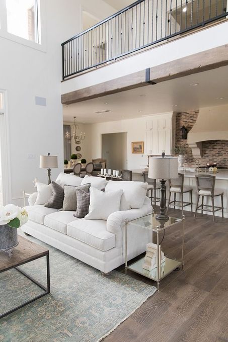 Bright open living room with white sofa, glass coffee table, and kitchen bar stools under a loft railing
