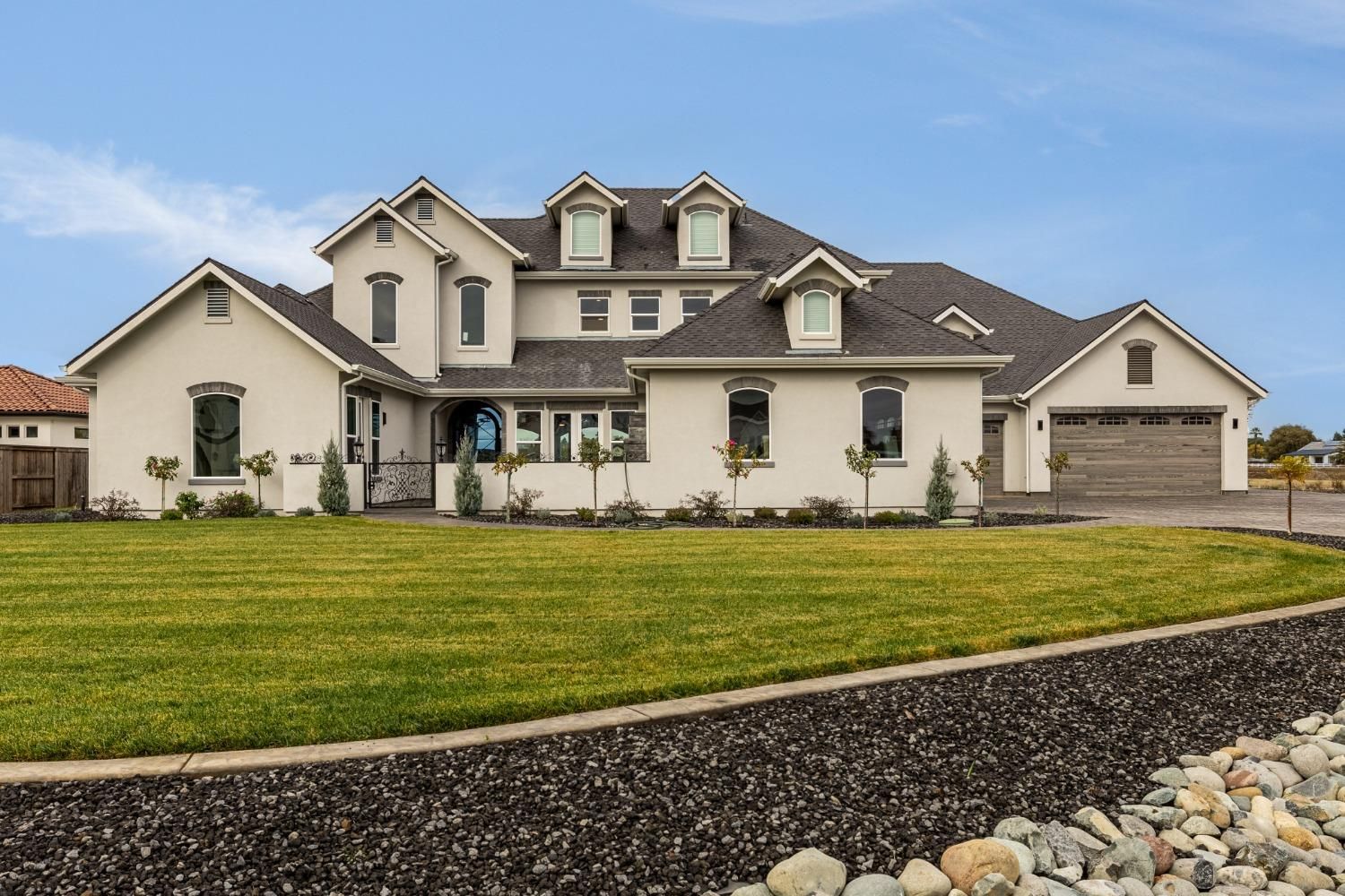 Large beige suburban house with a dark roof and manicured lawn under a blue sky
