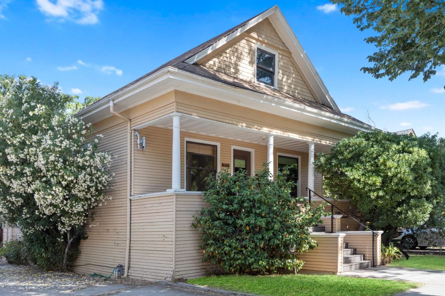 Small beige bungalow with front porch, steps, and shrubs under a clear blue sky