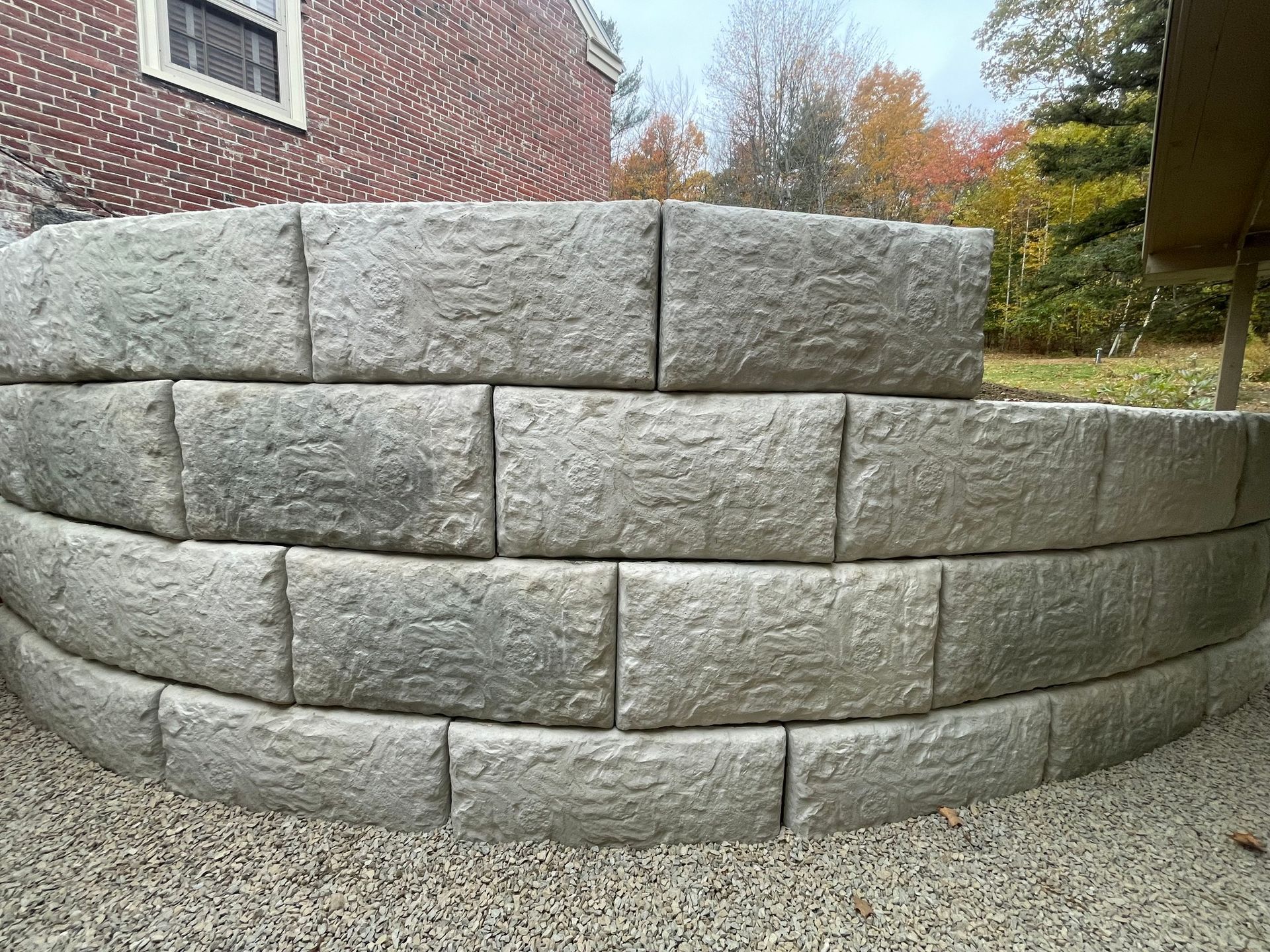 Curved stacked stone retaining wall beside a brick house in a yard, with autumn trees in the background