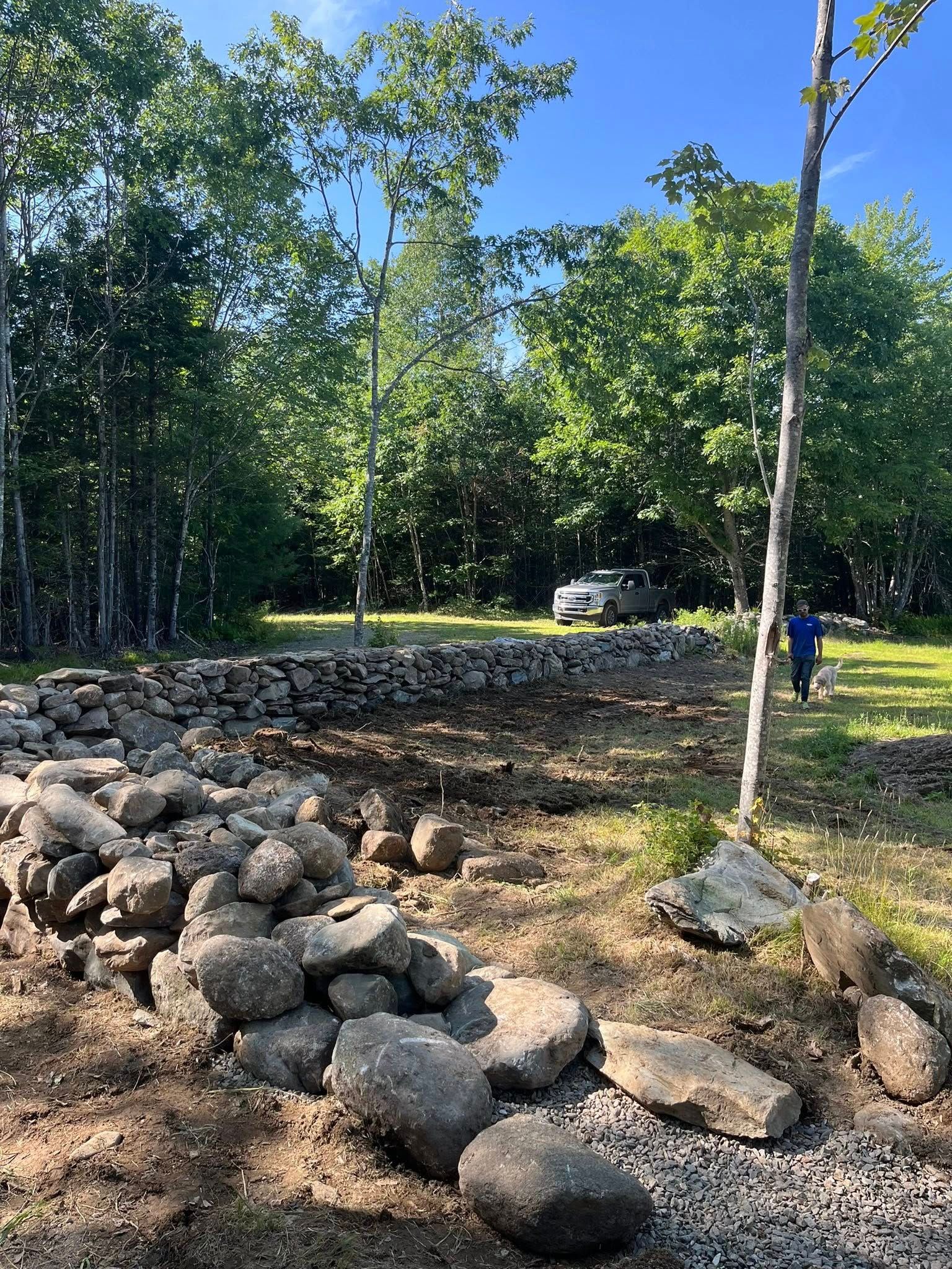 Rock-lined garden path with trees and a person in blue near a white car in the background