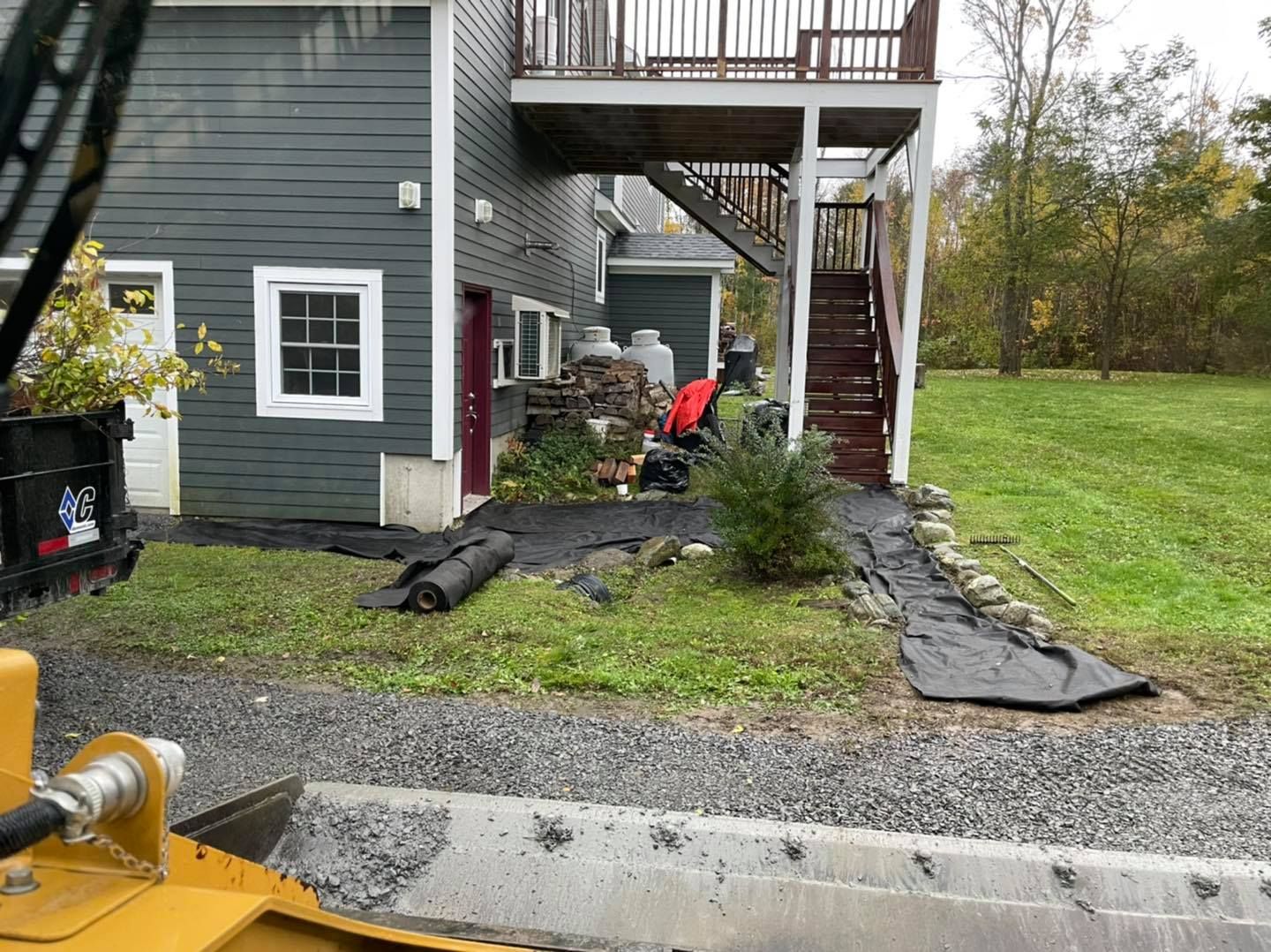 Backyard by a gray house with gravel path, tarps, and a worker under a raised deck