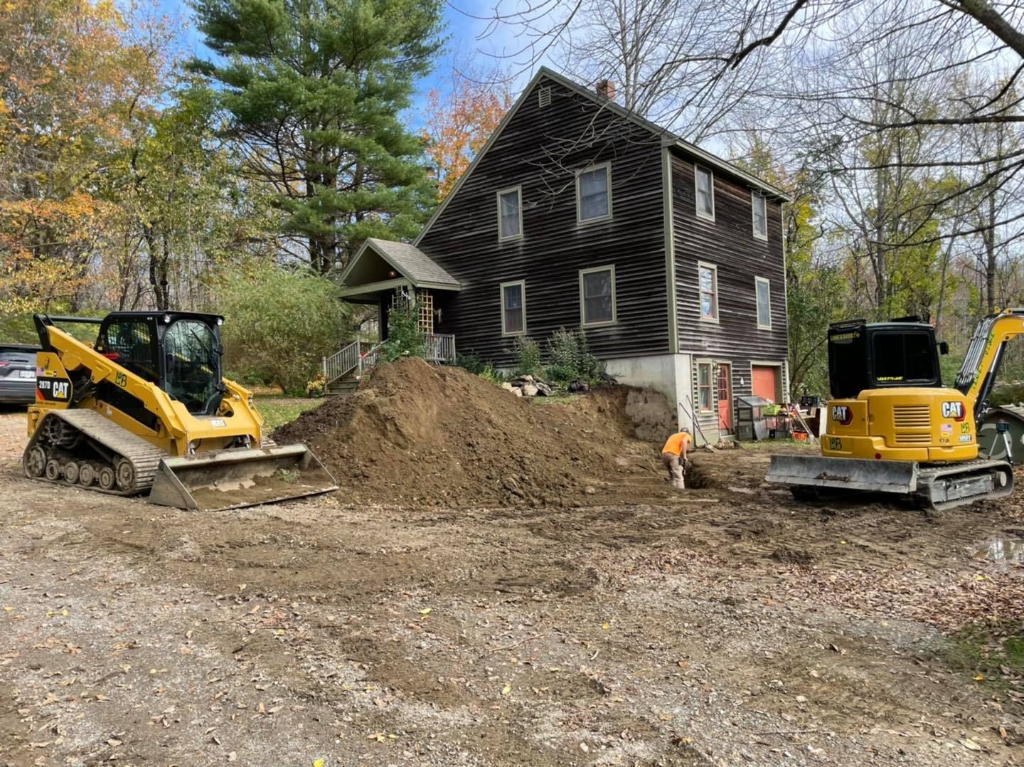 Construction equipment digging dirt beside a dark two-story house with trees in the background