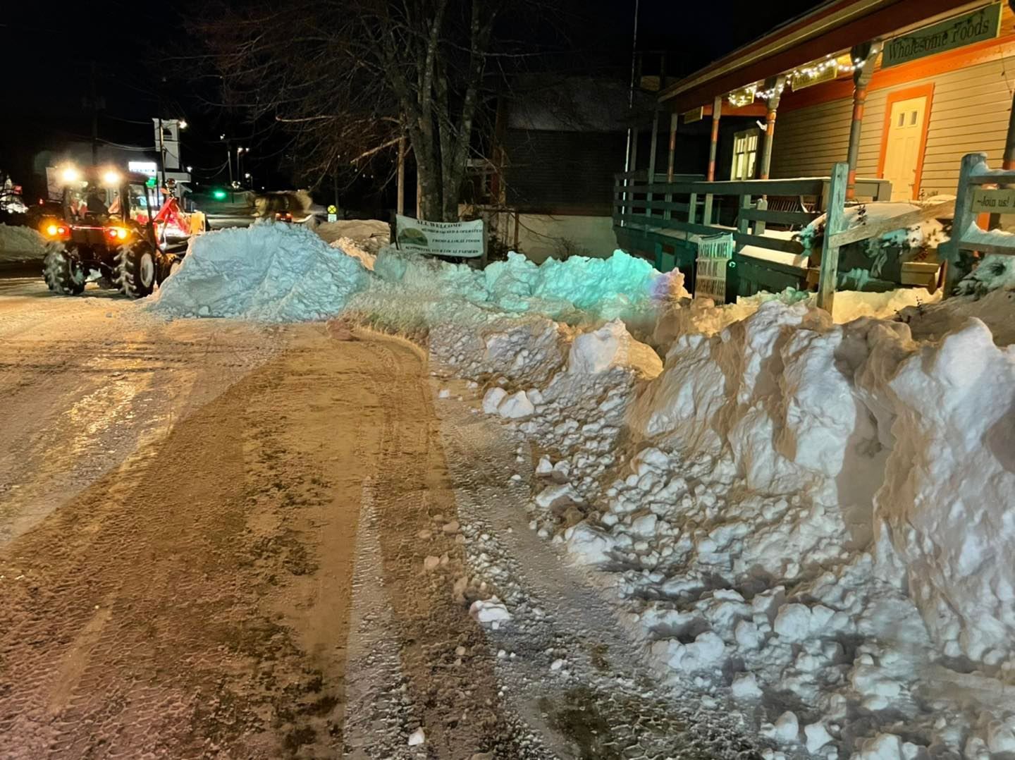 Snow-covered road at night with a lit cabin, snowbanks, and a vehicle plowing on the left