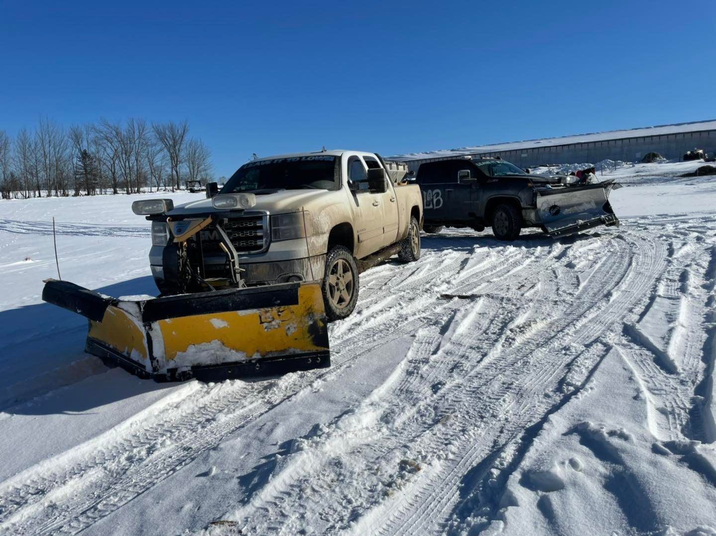 Snowplow pushing snow in front of a tan pickup truck on a snowy roadside near other vehicles