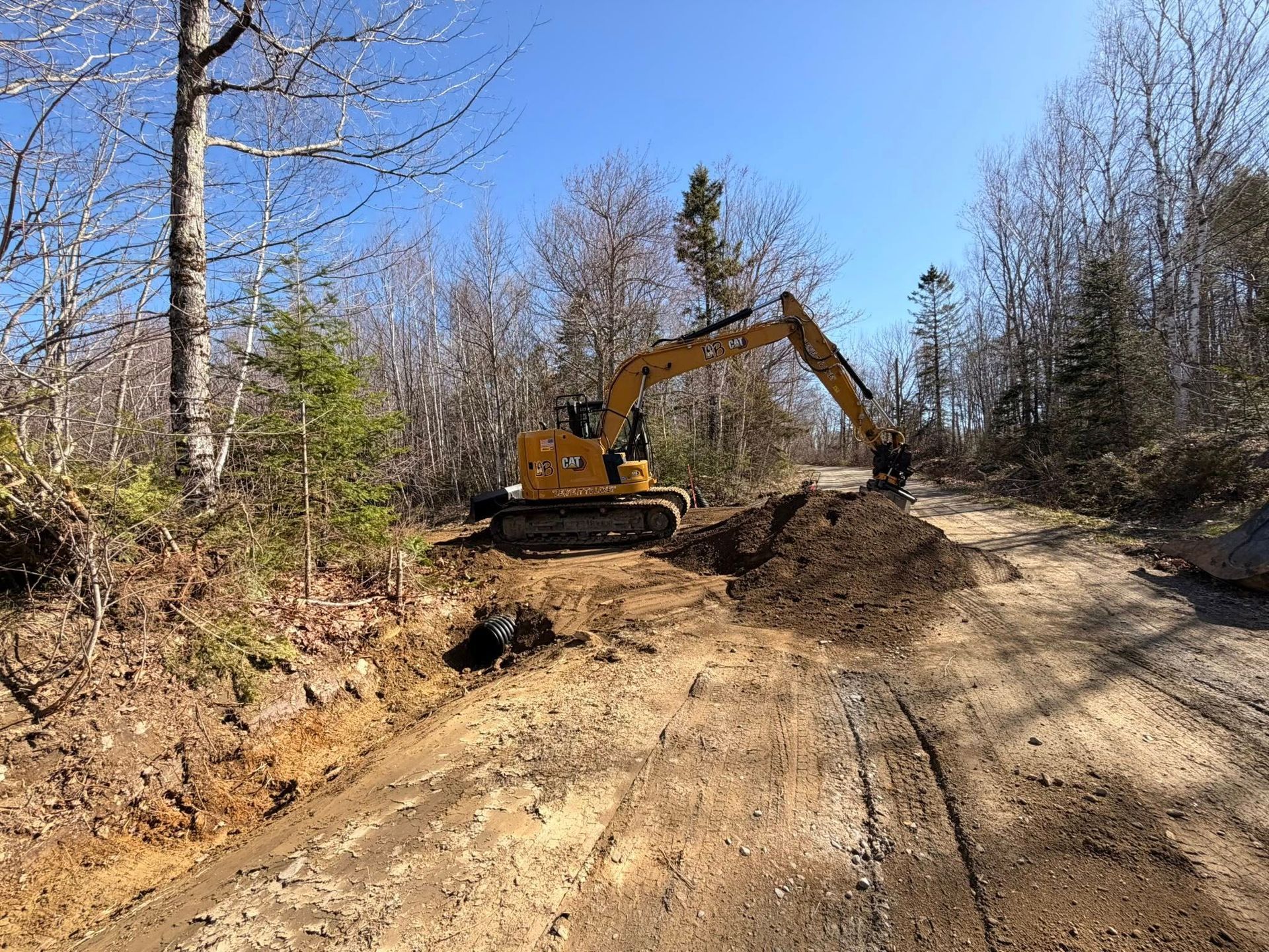 Excavator working on a dirt road in a wooded area under a clear blue sky