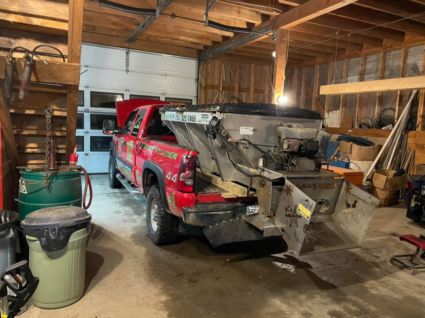 Red pickup truck inside a wooden garage, loaded with a metal front-end loader attachment.