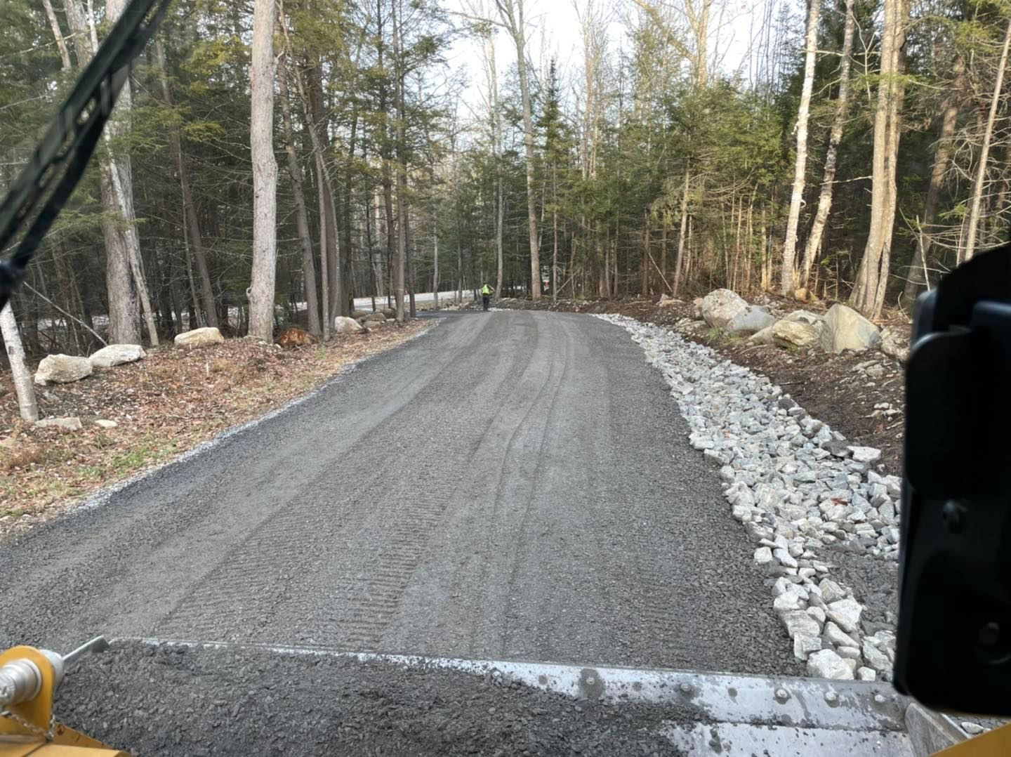 Gravel road under construction through a wooded area, lined with rocks and trees.