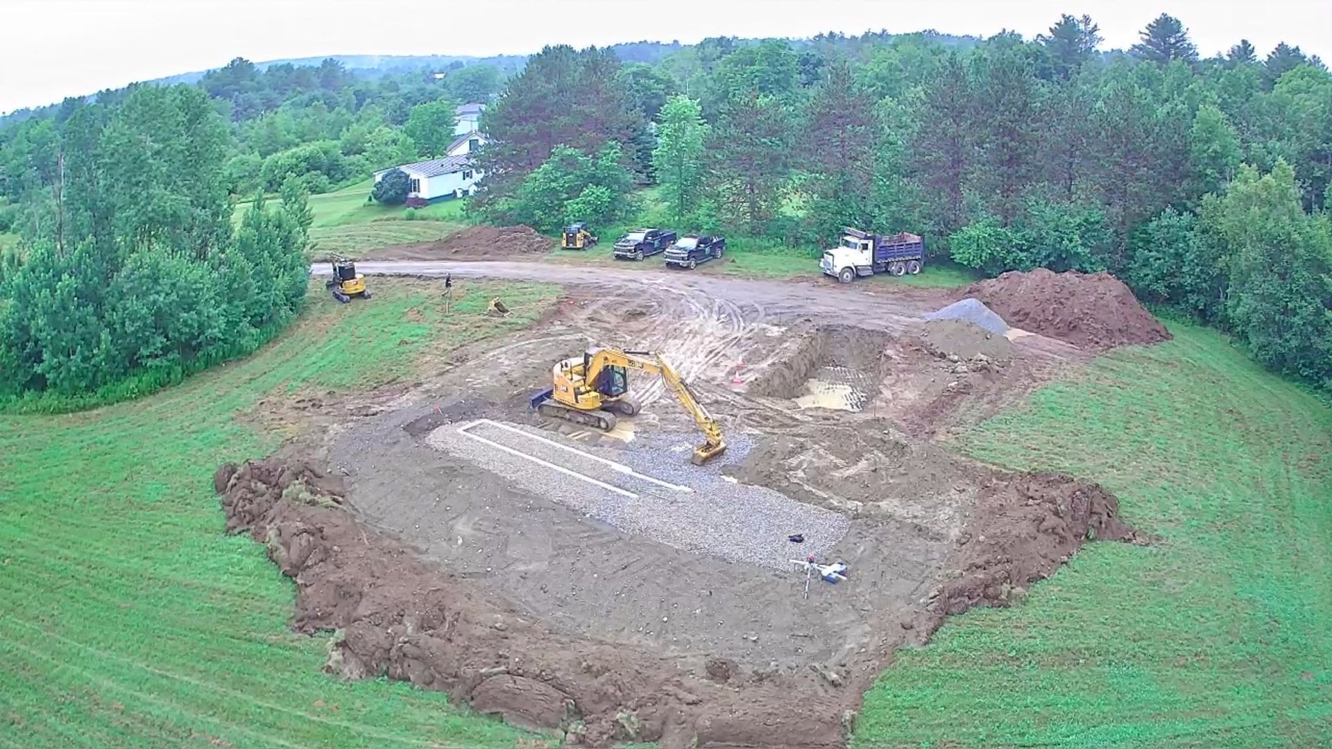 Aerial view of a construction site with an excavator clearing a large dirt area surrounded by green trees.