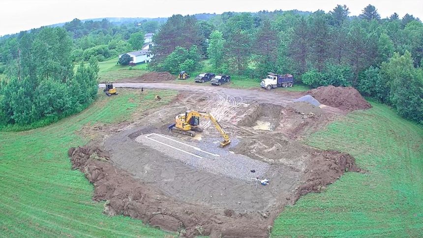 Excavator working at a large dirt construction site surrounded by green trees and grass