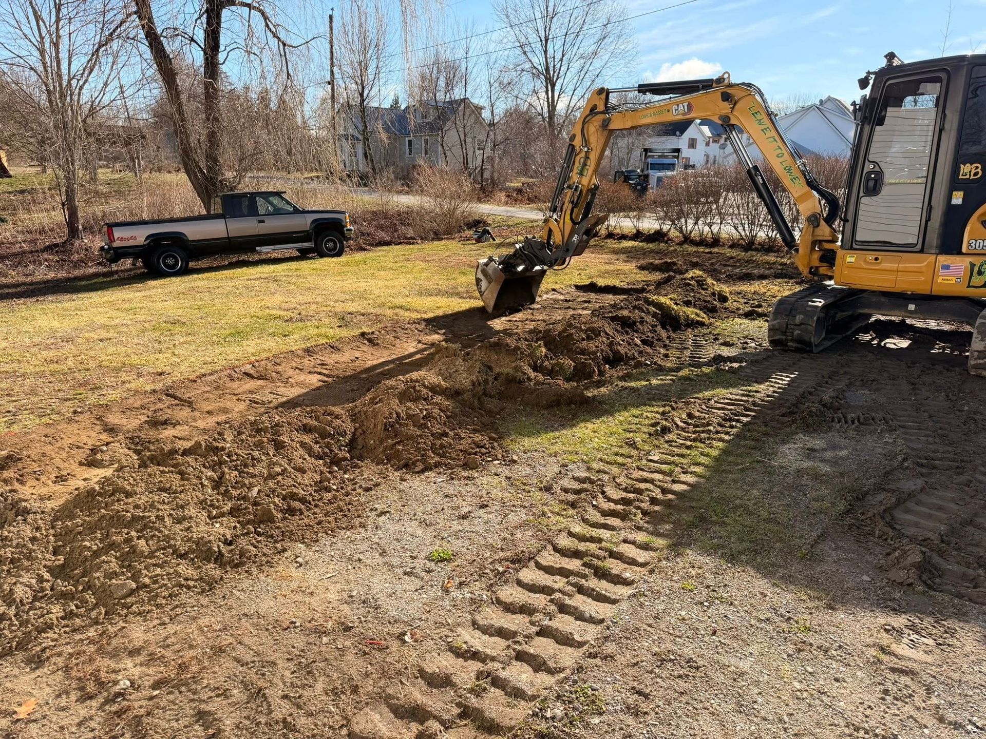 Excavator digging muddy ground beside a black pickup truck in a wooded lot.