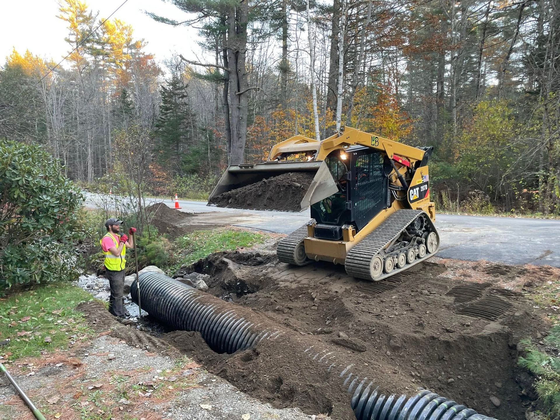 Skid steer excavator dumping soil into a trench while a worker stands nearby in a wooded area