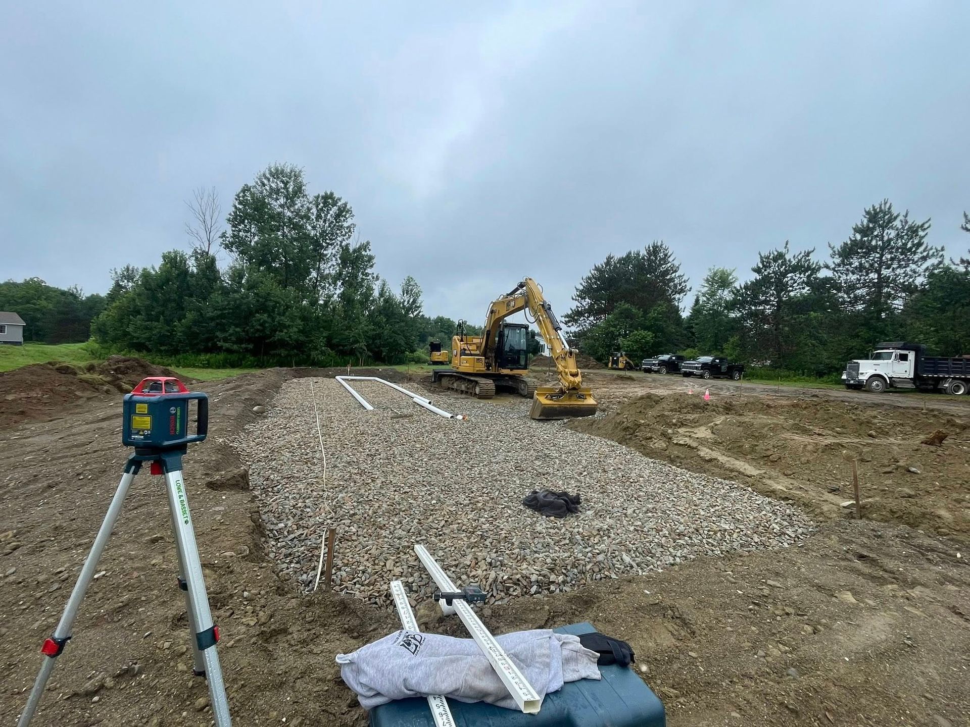 Construction site with excavator on gravel strip, survey tripod in foreground, and cloudy sky.