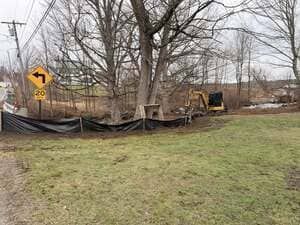 Yellow road sign beside a fenced construction area with a backhoe and bare trees beside a grassy field
