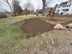 Freshly landscaped yard with a large dirt mound, rocks, and a backhoe near a house