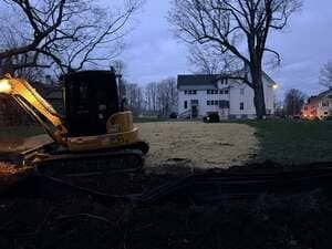 Excavator working at dusk in front of a white house and bare trees.