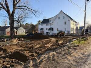 Construction site beside a white house with dirt, trenches, and a large tree by the road