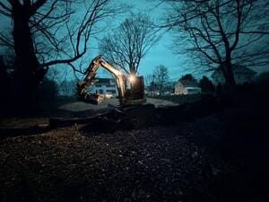 Excavator at dusk in a wooded construction site, with bare trees and a blue sky.