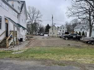 Residential street with houses, parked truck, and muddy vacant lot under cloudy sky