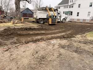 Construction site with excavated dirt, a dump truck, and a backhoe beside a white house.