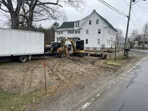 Construction equipment beside white house on muddy roadside, with truck and power lines nearby.