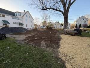Residential yard with fresh dirt, a large tree, and white houses in the background