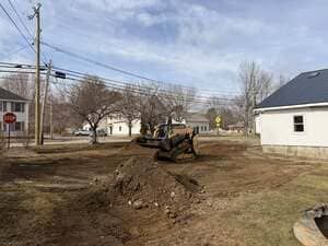 Construction vehicle digging a muddy trench on a residential street near houses and power lines