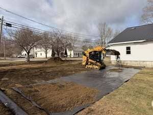 Backhoe smoothing soil in a yard beside a white house under a cloudy sky
