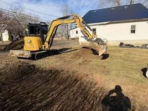 Yellow excavator grading a dirt yard beside a white house on a sunny day
