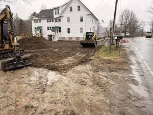 Construction site beside white house on a rainy street, with excavators and muddy earthmoving work