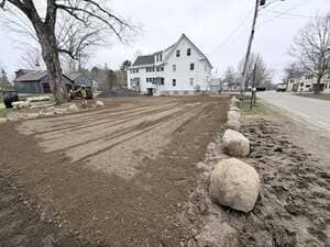 Freshly graded dirt lot beside a white house and roadside, bordered by large boulders and bare trees
