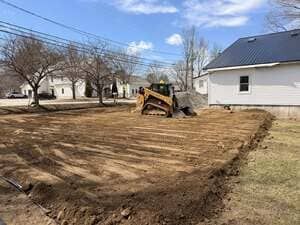 Bulldozer grading a dirt lot beside a white house under a partly cloudy sky