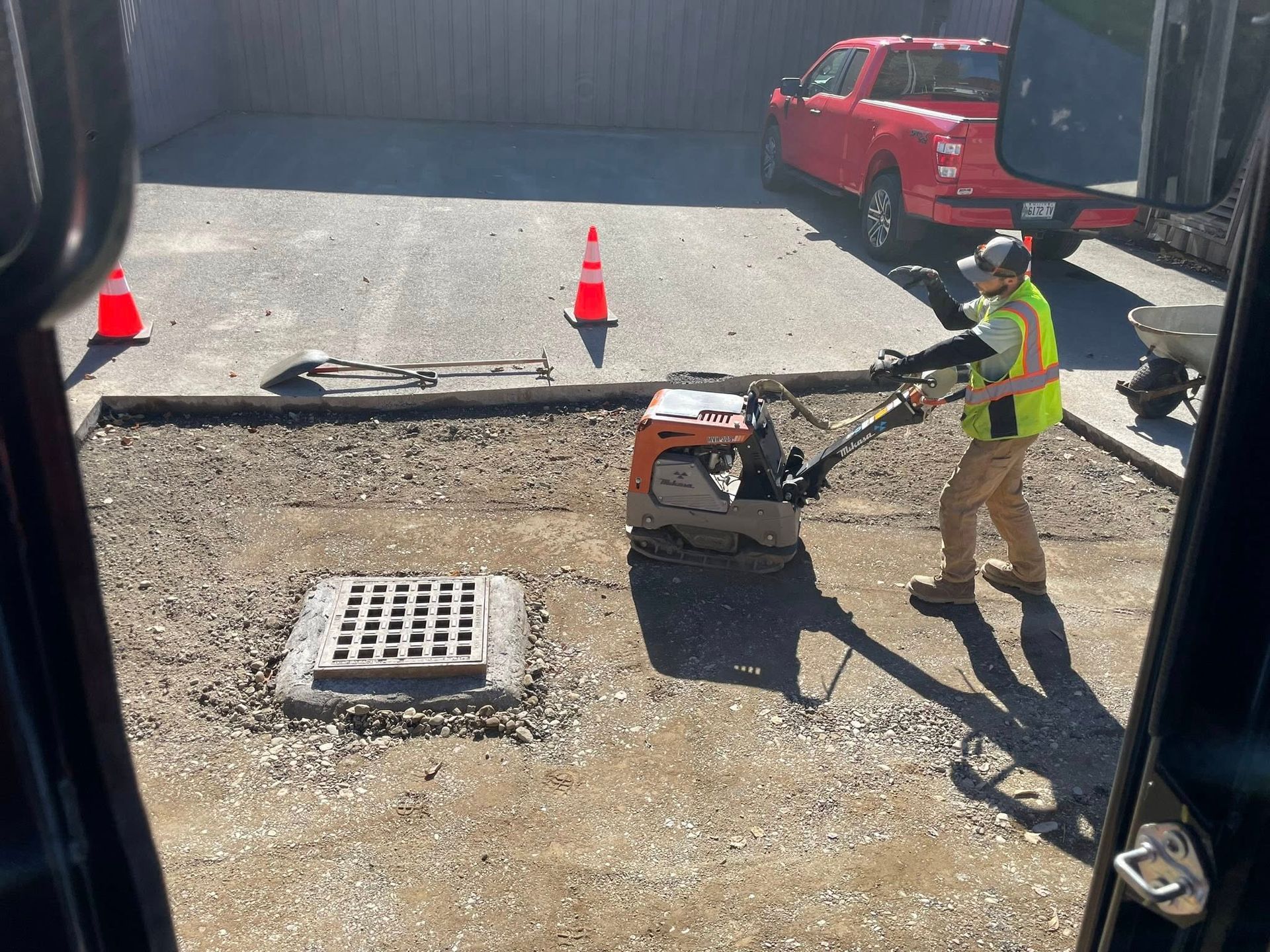 Worker in neon vest operating a compacting machine on a gravel lot near cones and a red truck