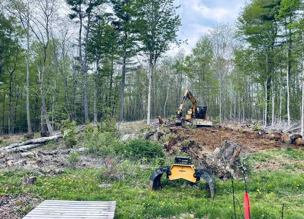 Excavator clearing a muddy forest clearing with birch trees and a yellow machine in the foreground