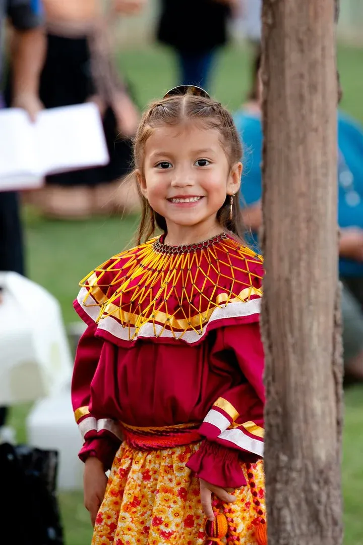 A little girl in a red and yellow dress is standing next to a tree.