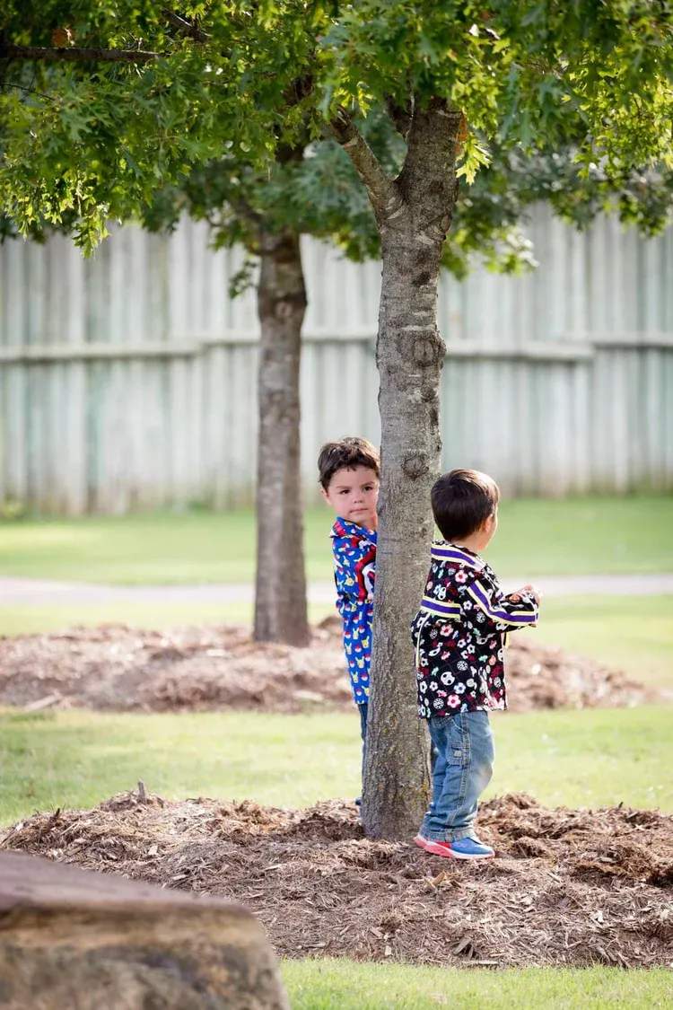 Two young boys are hiding behind a tree in a park.