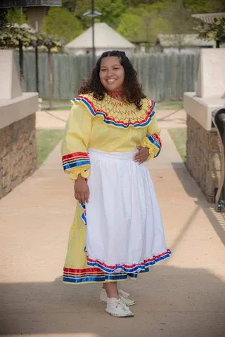 A woman in a yellow dress and white apron is standing on a sidewalk.