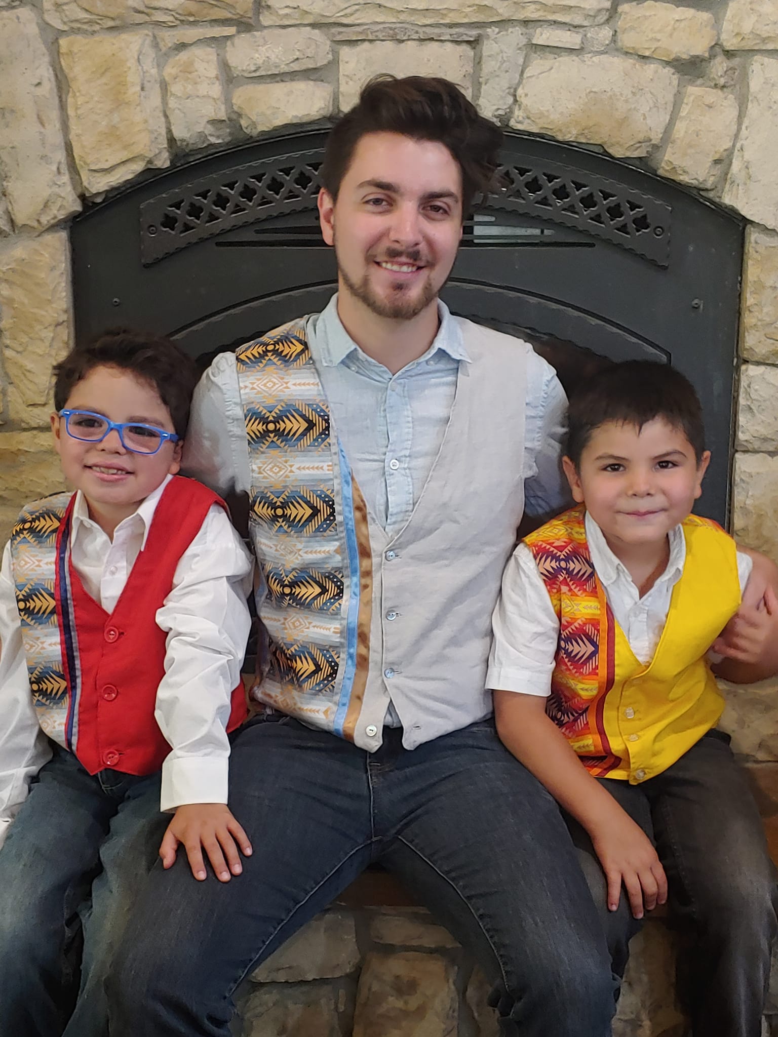 A man is sitting next to two young boys in front of a fireplace wearing vests.