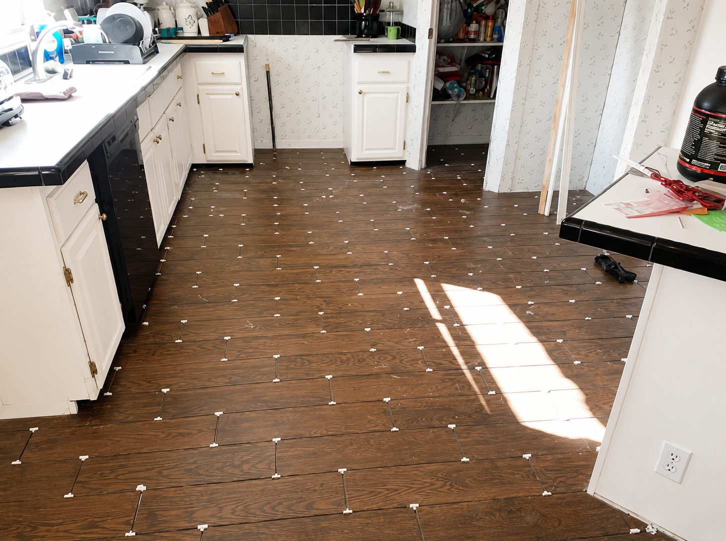 Sunlit kitchen with white cabinets, dark wood floor, and scattered white crumbs or debris.