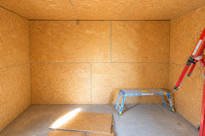Empty unfinished room with plywood walls, concrete floor, and a step stool near a ladder.